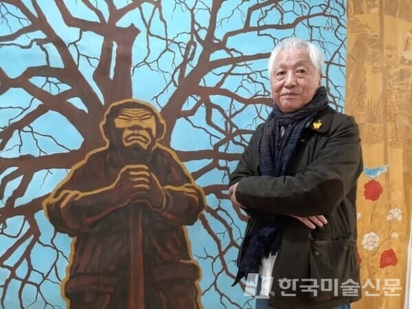 1994년 그린 대작 '말목장터 감나무 아래 아직도 서 있는&hellip;' 앞에 선 김정헌 작가. 2019년 김종영미술관 회고전 때 찍었다. 신간 '어쩌다 보니, 어쩔 수 없이'의 표지그림으로 쓰인 작품이다. 동학농민혁명 당시 첫 농민봉기가 일어난 전북 정읍 말목장터의 감나무를 배경으로 괭이 들고 선 농민의 굳건한 자태를 그렸다.